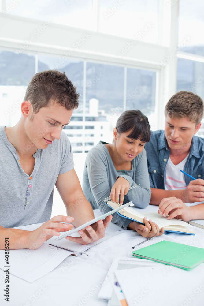 A group of students sit together using a tablet and a book to st A group of students sit together using a tablet and a book to st