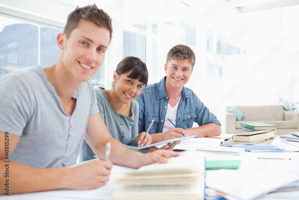 Three smiling students doing homework as they look into the came Three smiling students doing homework as they look into the came