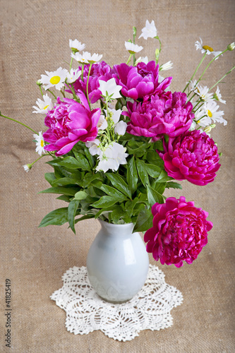 Bouquet of peonies and daisies in a white vase on a lace napkin © lenkusa