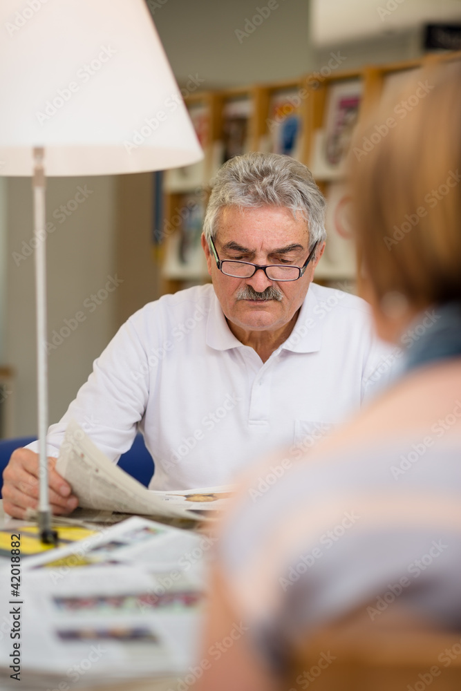 Senior man reading newspaper with wife in library Senior man reading newspaper with wife in library