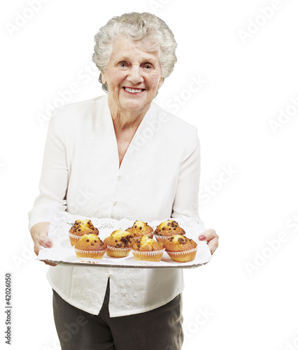 senior woman smiling and holding a tray with muffins against a w