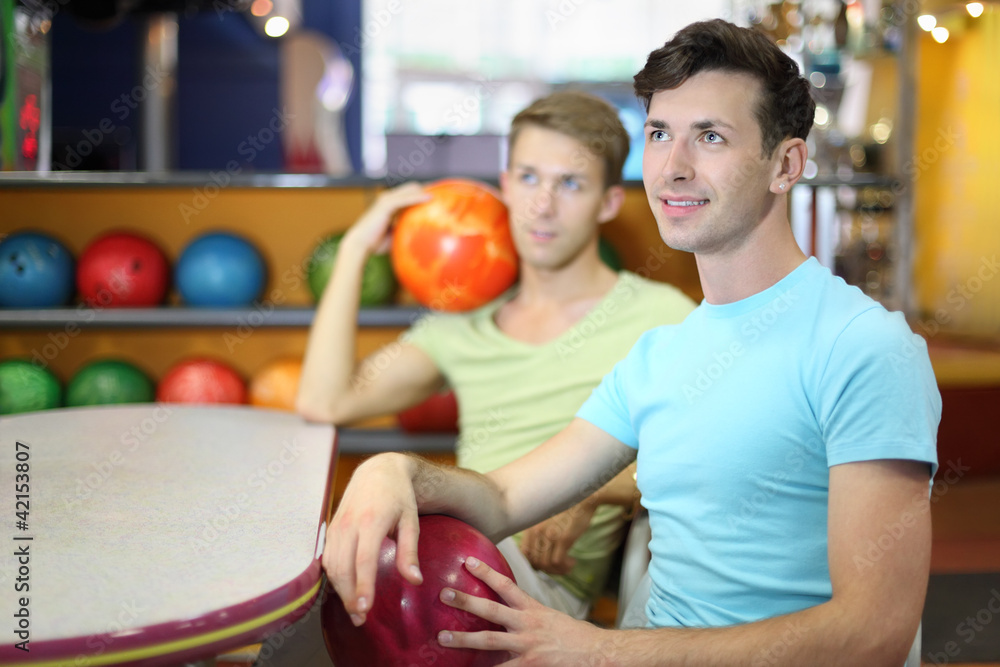 Fototapeta premium Two men sit at table in bowling, hold balls and look up