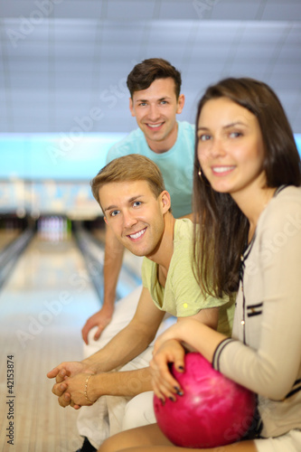 Two smiling men and woman with pink ball sit in bowling club