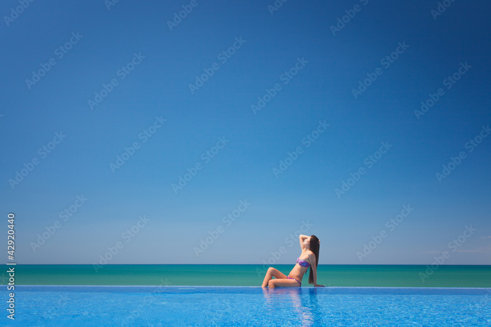 Beautiful woman sitting near infinity pool and looking to sea an