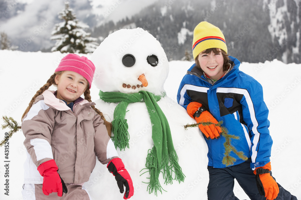 Two Young Children Building Snowman On Ski Holiday In Mountains Two Young Children Building Snowman On Ski Holiday In Mountains