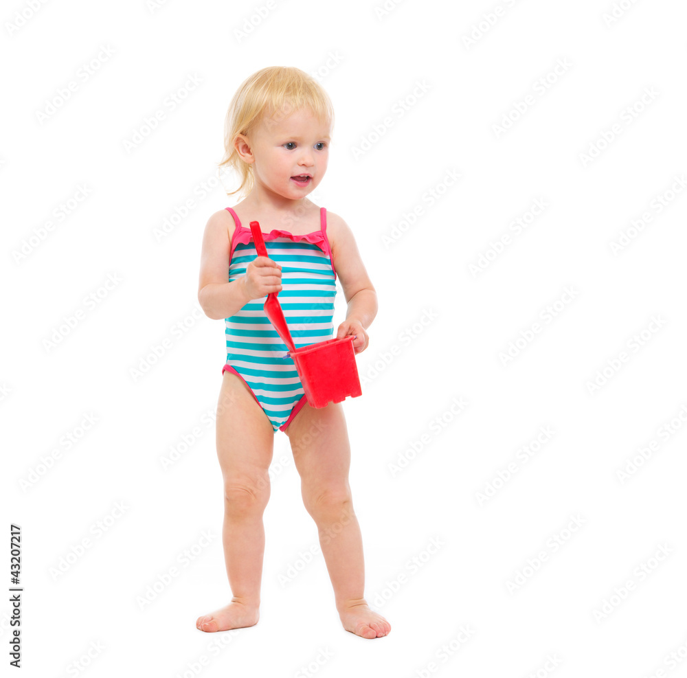 Baby girl in swimsuit holding bucket and shovel looking in corne Baby girl in swimsuit holding bucket and shovel looking in corne