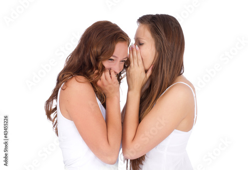 Two smiling young girlfriends talking a secret over white