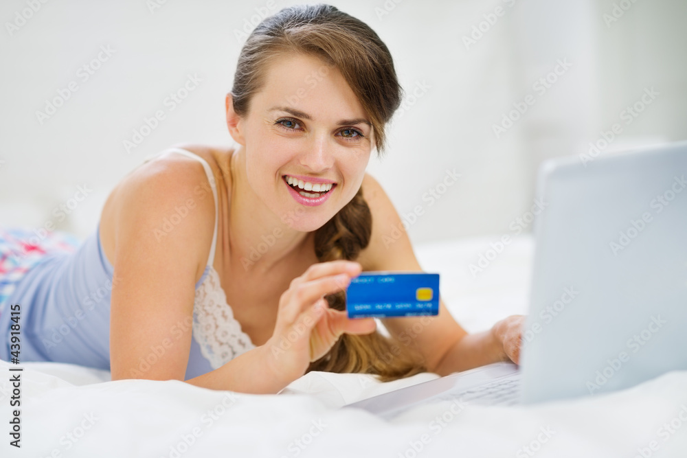 Happy young woman laying on bed with credit card and laptop Happy young woman laying on bed with credit card and laptop