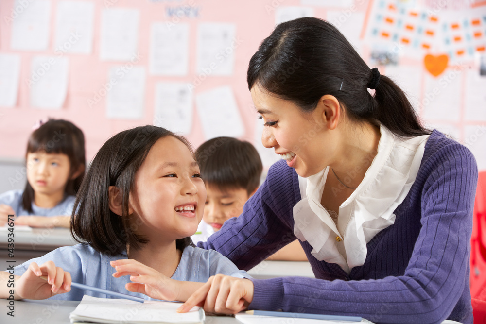 Teacher Helping Student Working At Desk In Chinese School Teacher Helping Student Working At Desk In Chinese School