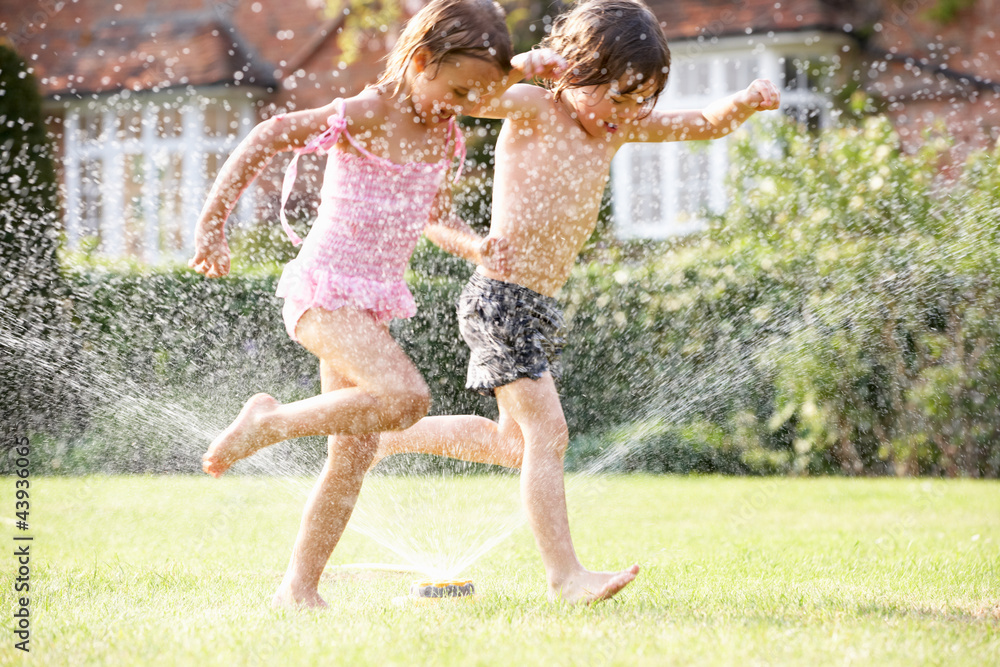 Two Children Running Through Garden Sprinkler - obrazy, fototapety, plakaty 