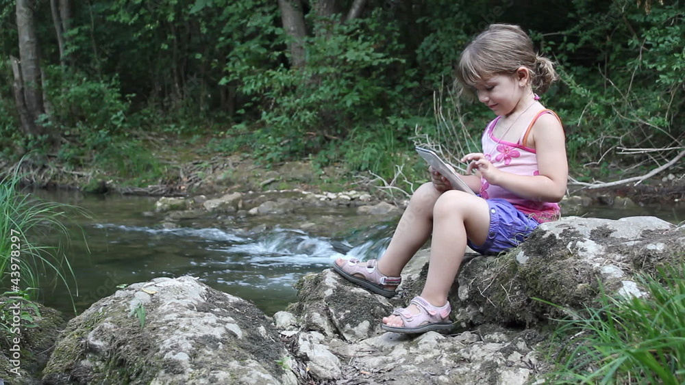 little girl sitting next to a stream and play with tablet little girl sitting next to a stream and play with tablet