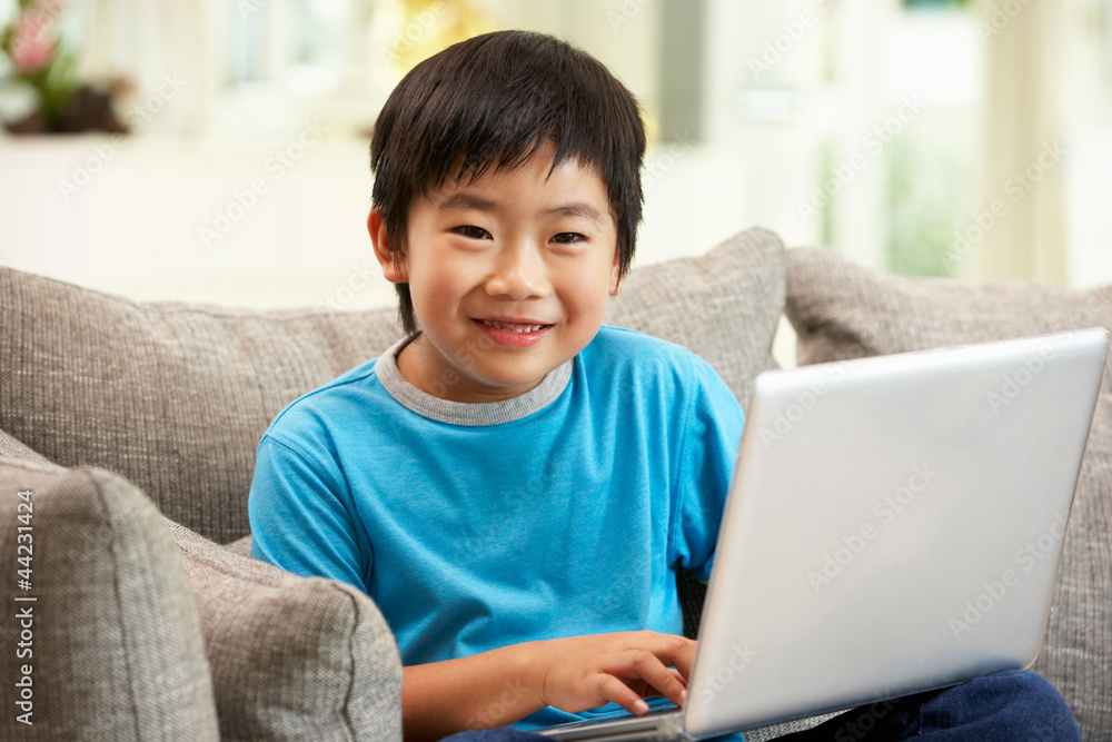 Young Chinese Boy Using Laptop Whilst Sitting On Sofa At Home Young Chinese Boy Using Laptop Whilst Sitting On Sofa At Home