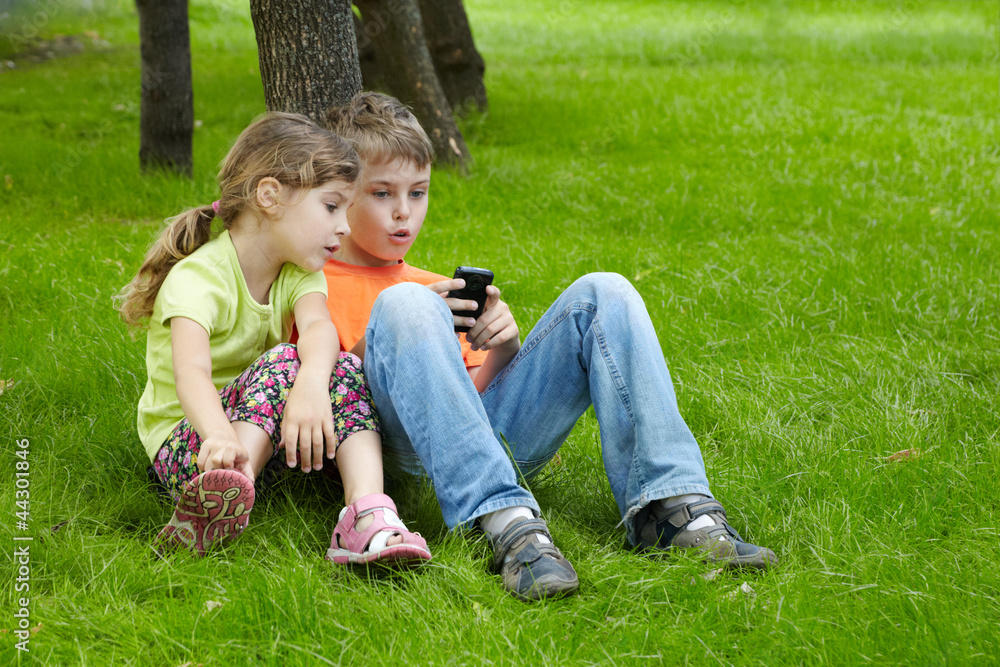Boy and his younger sister sit on grass under tree in park Boy and his younger sister sit on grass under tree in park