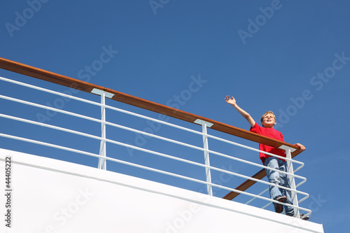Boy stands at railing on deck of ship, shouts and waves his hand