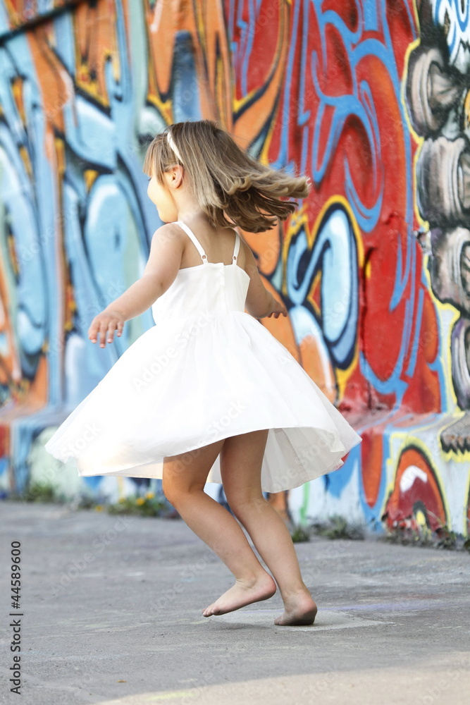 Kleines Mädchen in weißem Kleid tanzt vor Graffiti Wand