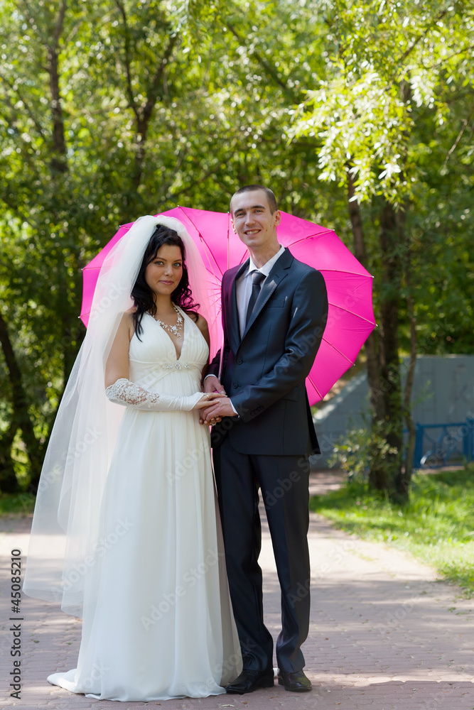Newly-married couple in a summer garden under a pink umbrella