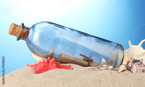 Glass bottle with note inside on sand, on blue background