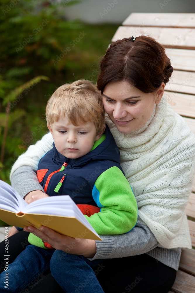 Young mother and toddler son reading book on bench outdoor Young mother and toddler son reading book on bench outdoor
