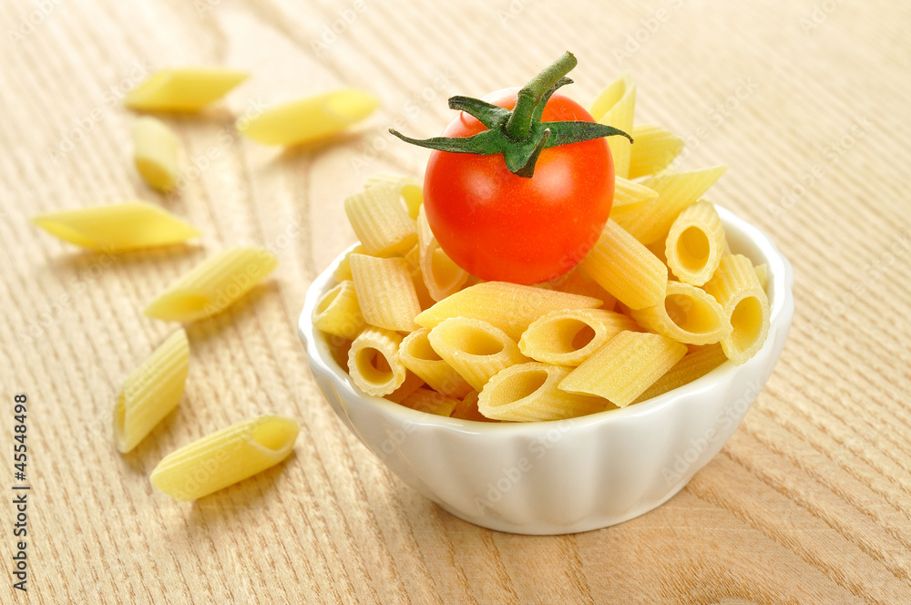 Several uncooked penne pasta and a cherry tomato in a small bowl