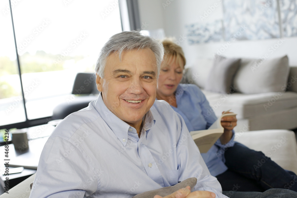 Obraz premium Smiling senior man sitting in couch, wife in background