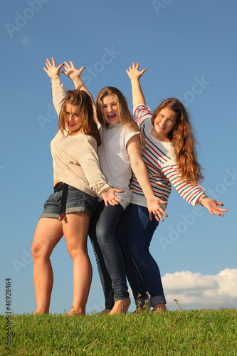 Three happy girls pose at green grass at background of blue sky.