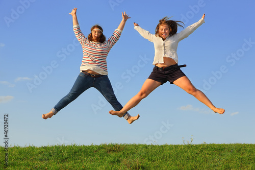 Two young fat girls jump at green grass at background of sky