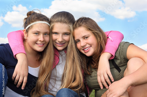 Three happy smiling girls hug at background of sky with clouds.
