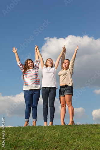 Three girls hold raised their hands and scream at green grass