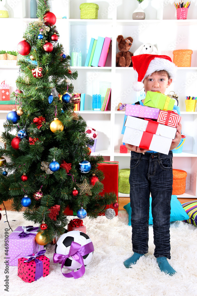 Little boy in Santa hat stands near Christmas tree with gifts Little boy in Santa hat stands near Christmas tree with gifts