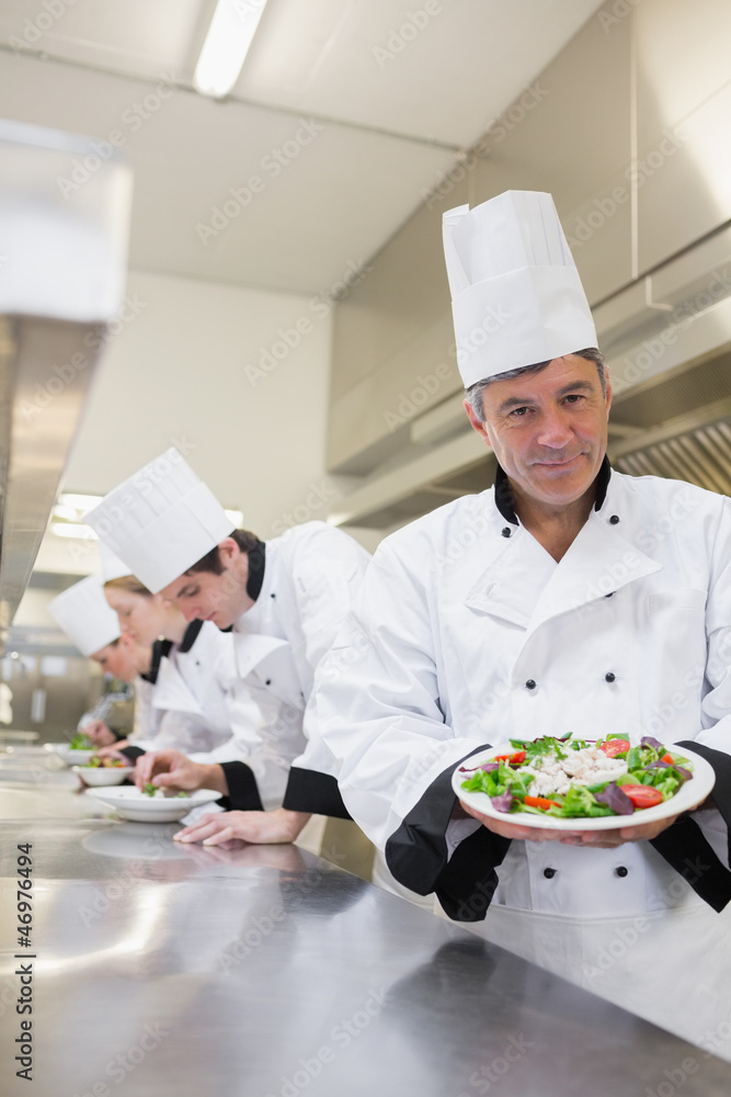 Cheerful chef showing his salad with workers in background Cheerful chef showing his salad with workers in background