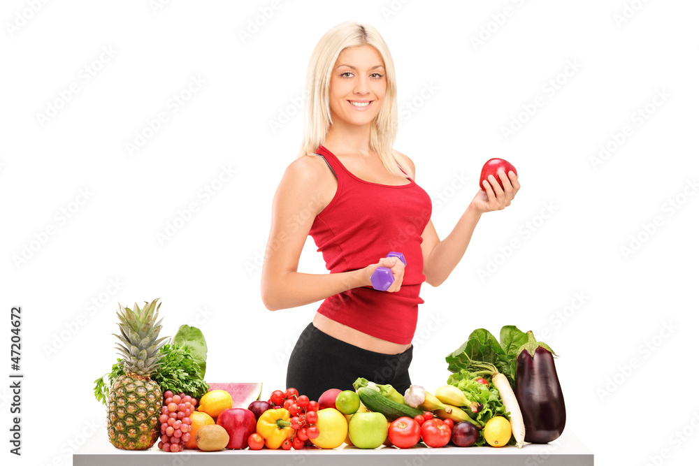 Female athlete holding a dumbbell and apple behind a pile of foo Female athlete holding a dumbbell and apple behind a pile of foo