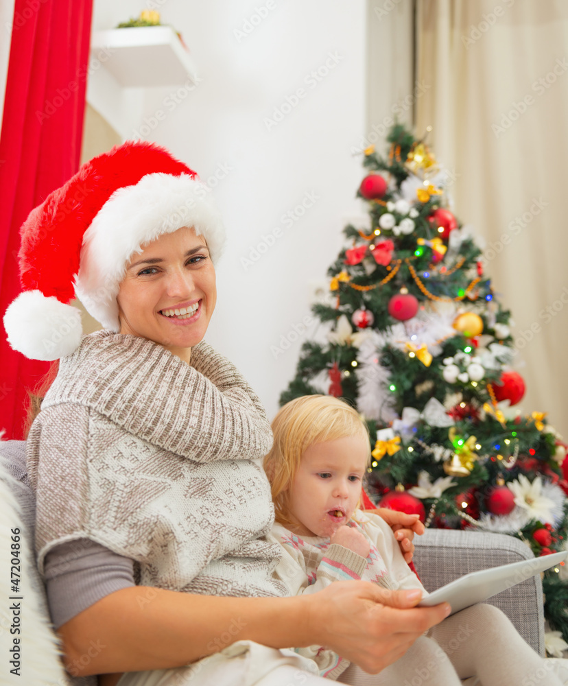Smiling mother and baby girl using tablet PC near Christmas tree Smiling mother and baby girl using tablet PC near Christmas tree