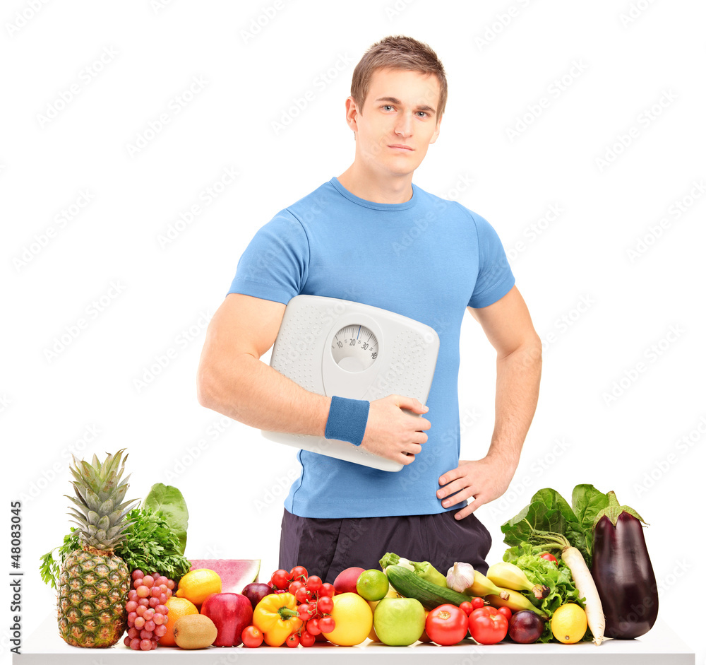 Male athlete holding a weight scale behind a table full of food Male athlete holding a weight scale behind a table full of food