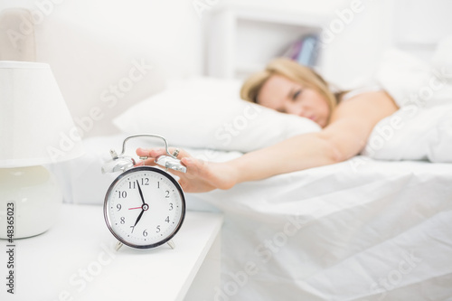 Irritated young woman in bed extending hand to alarm clock