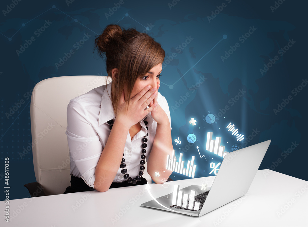 © ra2 studio - Pretty woman sitting at desk and typing on laptop with diagrams