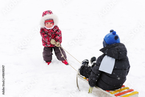 Cute kindergarten girl pulling her young brother on sled