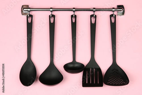 Black kitchen utensils on silver hooks, on pink background