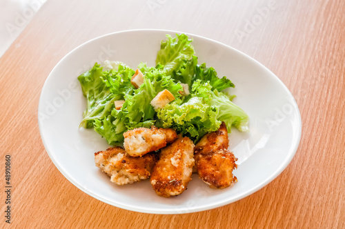 A simple white ceramic plate of chicken with some green salad leaves on a wooden table