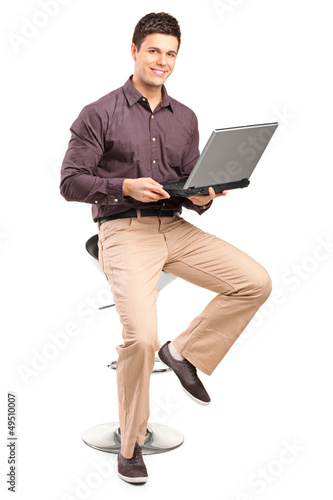 A smiling man sitiing on a high chair and working on a laptop