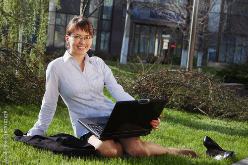 Woman lying on grass with laptop with business building on back