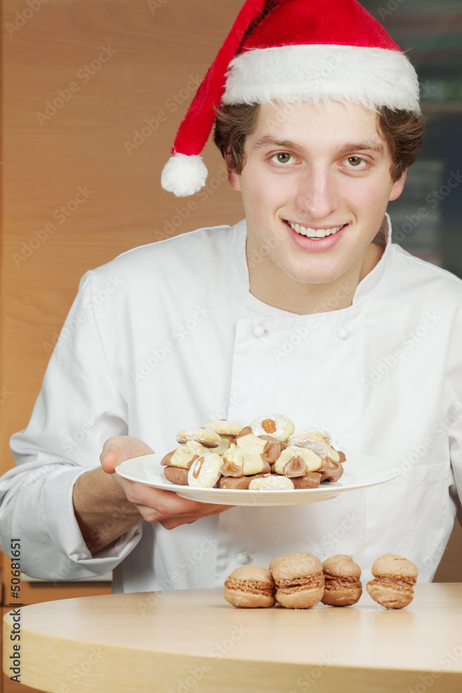 Smiling young confectioner in santa hat holds plate with cookies Smiling young confectioner in santa hat holds plate with cookies