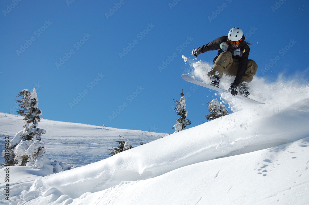 Snowboarder in action on a sunny winter day.  High jump from the hill followed by snow powder. Performs indy grab one hand. Blue sky and white snowy mountains on background. Extreme winter sports.