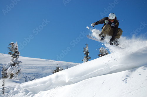 Snowboarder in action on a sunny winter day. High jump from the hill followed by snow powder. Performs indy grab one hand. Blue sky and white snowy mountains on background. Extreme winter sports.