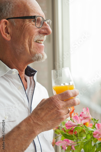 Senior man with glasses drinking orange juice at window with flo