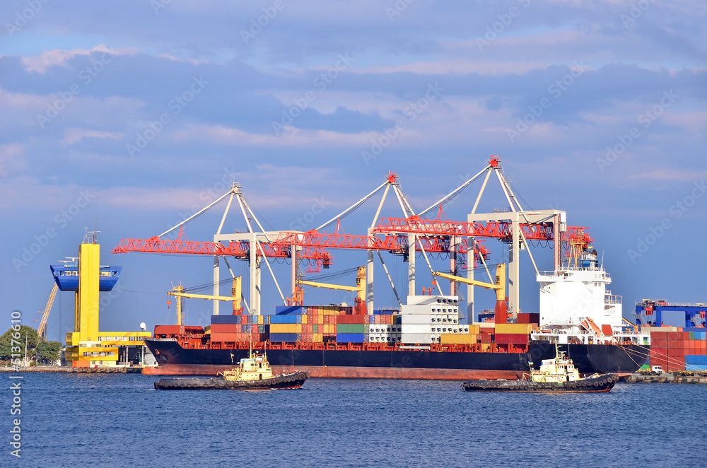 Container stack, tugboat and ship under crane bridge