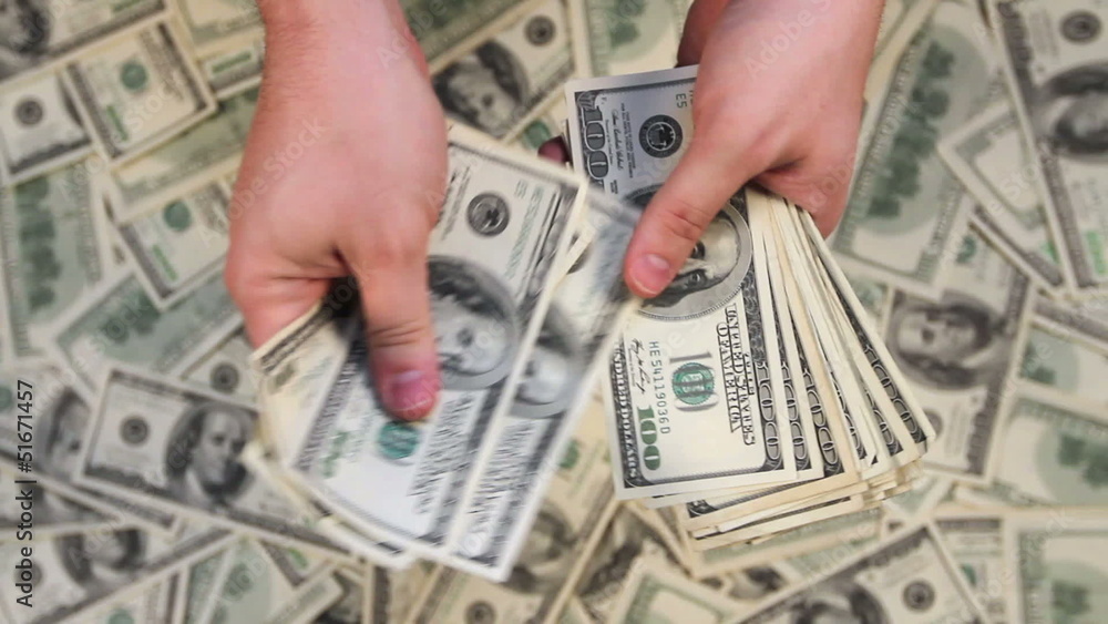 Looking down at hands counting a stack of hundred dollar bills with money in the background.
