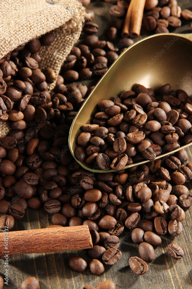 Fototapeta premium Roasted coffee beans in burlap sack with brass scoop and cinnamon sticks on dark wooden table, the rich brown tones of the coffee and wood create a warm, inviting atmosphere