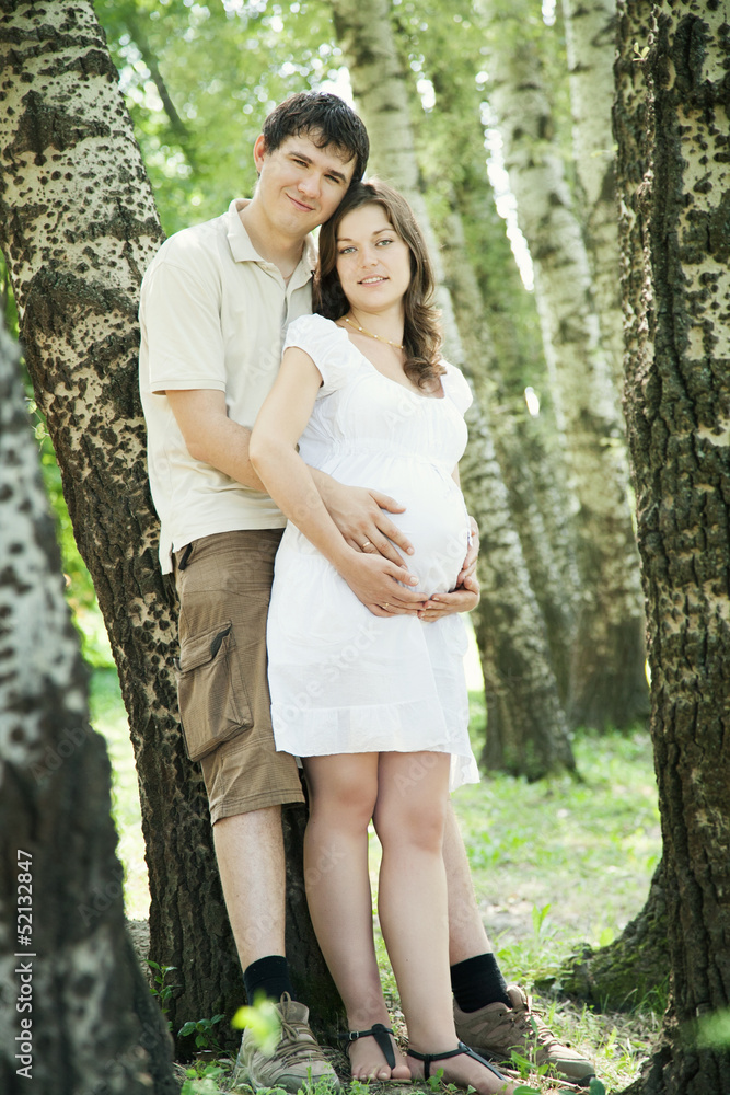 Fototapeta premium Pregnant wife and her husband in park leaning on trees