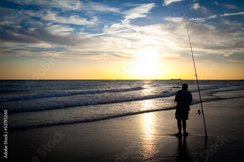 Fisherman silhouette while prepares his rod at the seashore at sunset