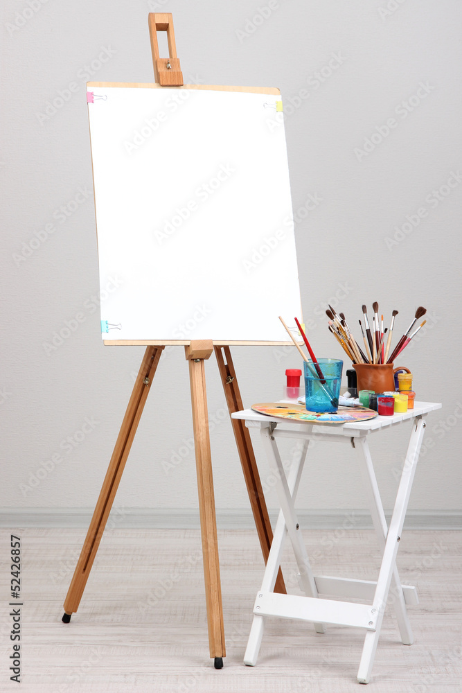 Wooden easel with clean paper and art supplies in room - obrazy, fototapety, plakaty 